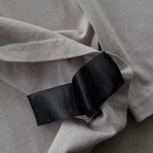 May include: A light beige t-shirt featuring a black fabric care label attached to the inner seam. The label displays various laundry instruction symbols and printed text detailing garment care, including washing instructions and material information.