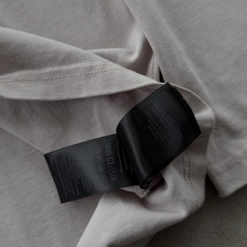 May include: A light beige t-shirt featuring a black fabric care label attached to the inner seam. The label displays various laundry instruction symbols and printed text detailing garment care, including washing instructions and material information.