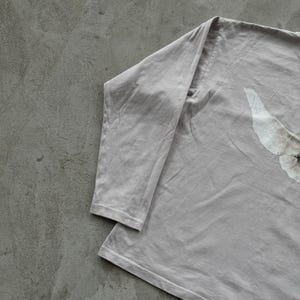 May include: A long-sleeve shirt in a muted light gray color. The garment features a partial graphic print on the right side, showing a white, textured wing-like shape with a small dark detail at the center of the design.