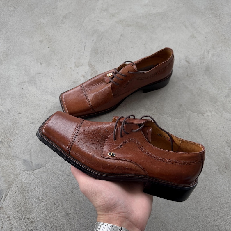 Puede incluir: Un par de zapatos oxford de cuero marr&oacute;n con punteras cuadradas y un detalle de costura decorativa. Los zapatos tienen un cierre de cordones y un logotipo dorado en el lateral.