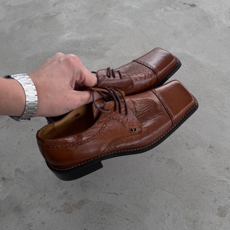 Puede incluir: Un par de zapatos de vestir de cuero marr&oacute;n con punteras cuadradas y un patr&oacute;n de costuras decorativas. Los zapatos tienen una suela de goma negra y una letra "V" dorada en relieve en el lateral.
