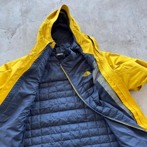 May include: A yellow hooded jacket with a navy blue quilted interior. The jacket has a zipper closure and the North Face logo on the chest. The jacket is open, revealing the interior lining.