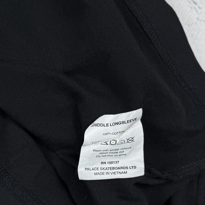 May include: A white tag with black text attached to a black long-sleeve shirt. The tag reads "GRIDDLE LONGSLEEVE", "100% COTTON", "Wash with similar colours", "Wash inside out", "Do not iron on print", "RN 150137", "PALACE SKATEBOARDS LTD", and "MADE IN VIETNAM".