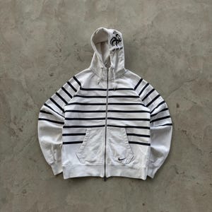 May include: White zip-up hoodie with navy blue horizontal stripes. Features a hood with a black rooster emblem and "FFF" text. Includes a front pocket and a small Nike logo. A casual, sporty garment.