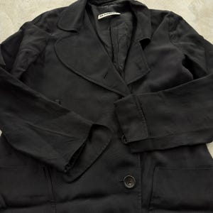 May include: A black blazer with long sleeves, a notched lapel, and two visible buttons on the front. The garment features a Jil Sander brand label on the inside collar and includes two large patch pockets on the lower front section of the jacket.