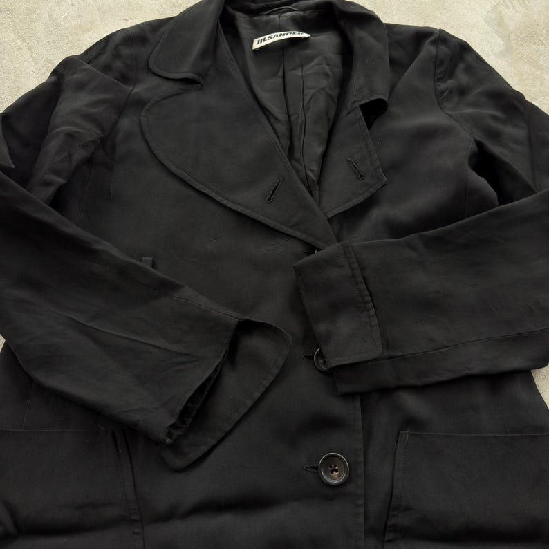 May include: A black blazer with long sleeves, a notched lapel, and two visible buttons on the front. The garment features a Jil Sander brand label on the inside collar and includes two large patch pockets on the lower front section of the jacket.