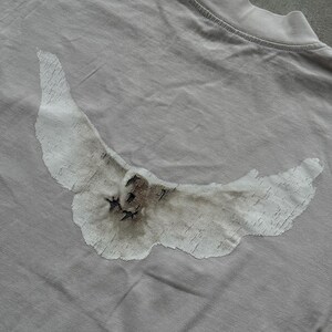 May include: A light gray t-shirt featuring a large, distressed white graphic of a bird in flight across the back. The bird design has a textured, weathered appearance with dark brown and black shading details visible on the wings and body.