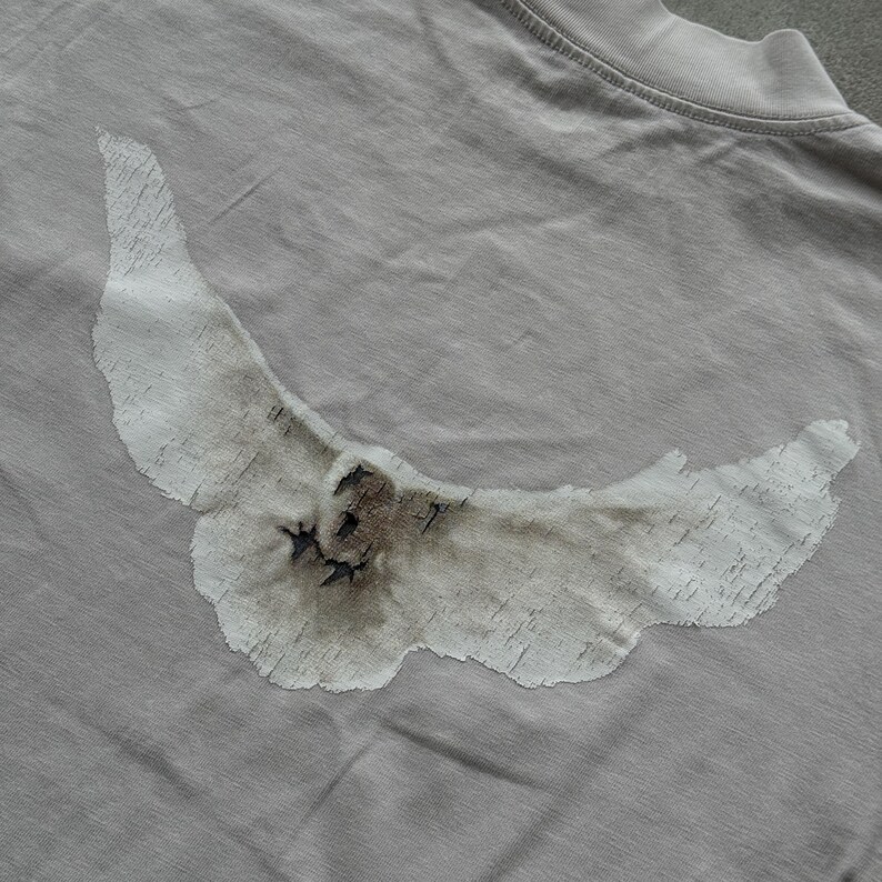 May include: A light gray t-shirt featuring a large, distressed white graphic of a bird in flight across the back. The bird design has a textured, weathered appearance with dark brown and black shading details visible on the wings and body.
