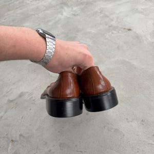 Puede incluir: Un par de zapatos de vestir de cuero marr&oacute;n con suelas negras. Los zapatos tienen un dise&ntilde;o de cordones y una punta ligeramente puntiaguda.