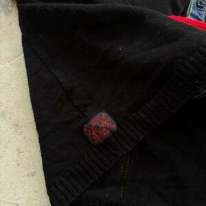 May include: Close-up of a black knitted sweater with a ribbed hem. A small, rectangular red patch with a circular design is sewn on the sleeve. The sweater is layered over a denim shirt with a red accent.