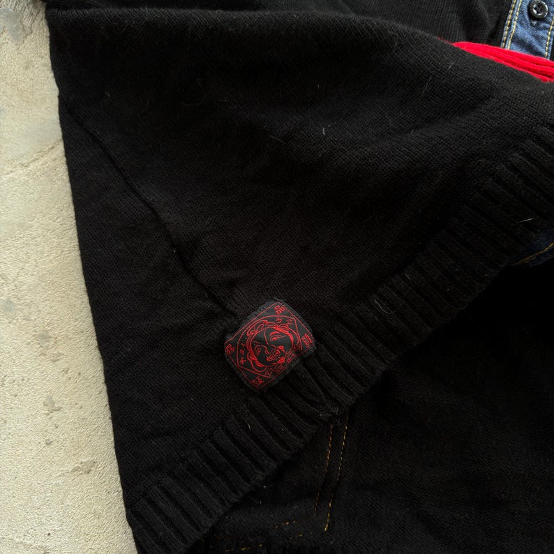 May include: Close-up of a black knitted sweater with a ribbed hem. A small, rectangular red patch with a circular design is sewn on the sleeve. The sweater is layered over a denim shirt with a red accent.