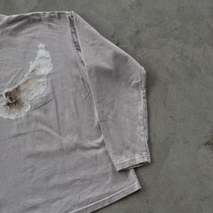 May include: A long-sleeve shirt in a muted light gray color featuring a distressed, textured white graphic print on the left side. The garment has a crew neckline, long sleeves with stitched cuffs, and a soft, worn-in fabric appearance throughout.