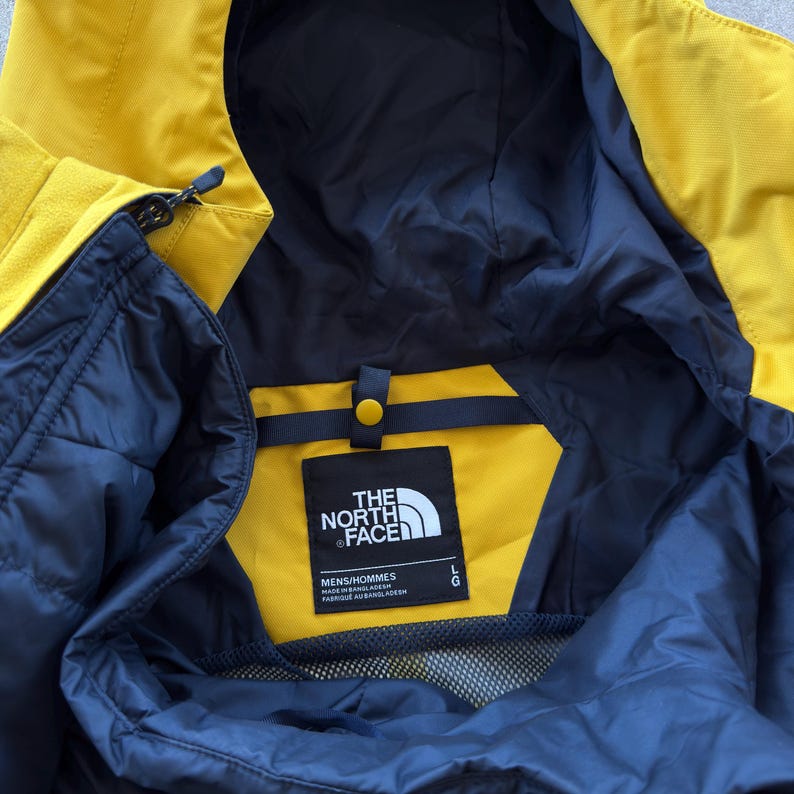 May include: Close-up of a yellow and navy blue jacket. The jacket has a hood and a zipper. The North Face logo is visible on a black tag. The jacket is labeled "MENS/HOMMES" and "L".