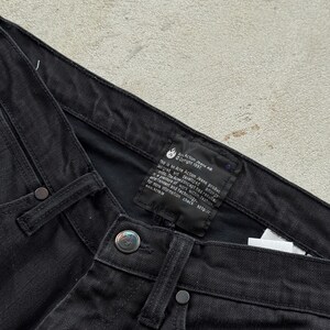 May include: Close-up of black denim jeans. The jeans have a button closure and rivets. A black label with white text is visible, including the text "Am Action Jeans AB Copyright 1997". The jeans are made of a sturdy fabric.