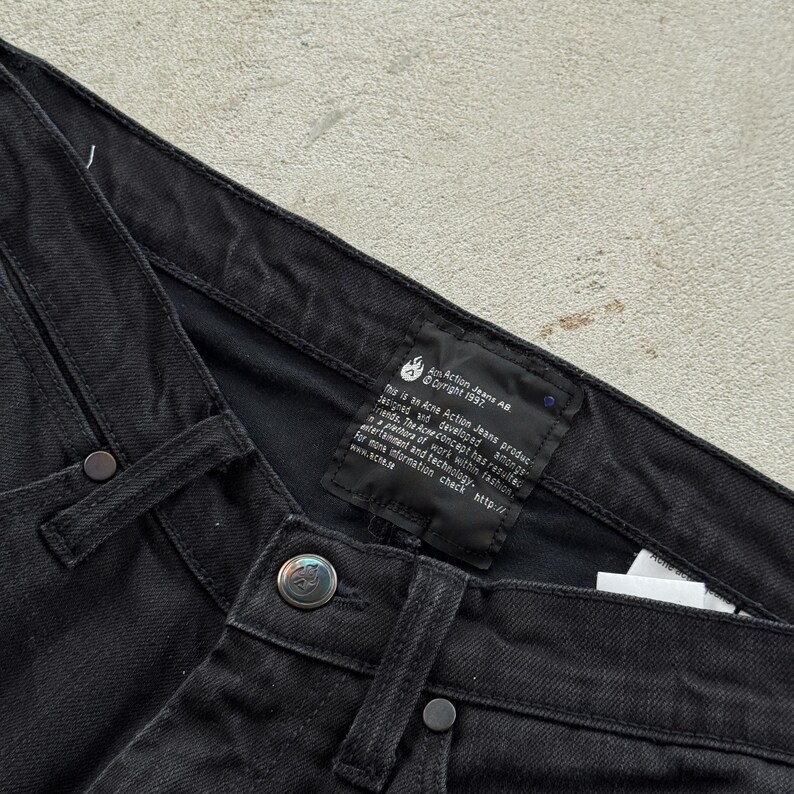 May include: Close-up of black denim jeans. The jeans have a button closure and rivets. A black label with white text is visible, including the text "Am Action Jeans AB Copyright 1997". The jeans are made of a sturdy fabric.