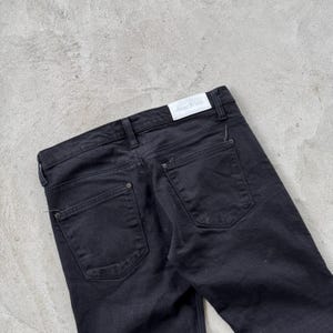 May include: Black denim jeans with a classic five-pocket design. The jeans feature a button and zipper closure, belt loops, and the brand name "Acne Jeans" on a white label. The back pockets have a simple, clean design.