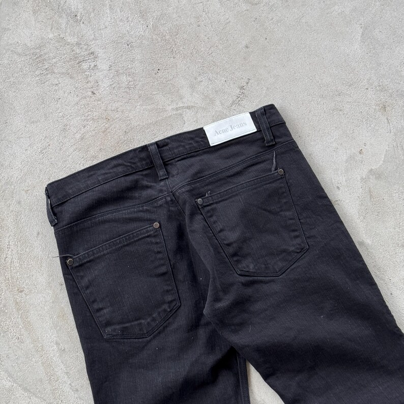 May include: Black denim jeans with a classic five-pocket design. The jeans feature a button and zipper closure, belt loops, and the brand name "Acne Jeans" on a white label. The back pockets have a simple, clean design.