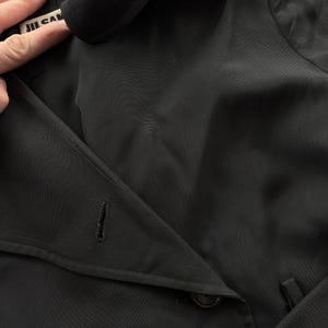 May include: A black jacket featuring a wide lapel, a single visible buttonhole, and a dark button closure. The interior collar tag displays the text JIL SANDER in black lettering on a white background, indicating the brand of this garment.