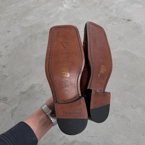 Puede incluir: Un par de zapatos de cuero marr&oacute;n con suela negra. Los zapatos tienen un logotipo V y el texto "Valentino" en la parte posterior. Los zapatos son talla 250.
