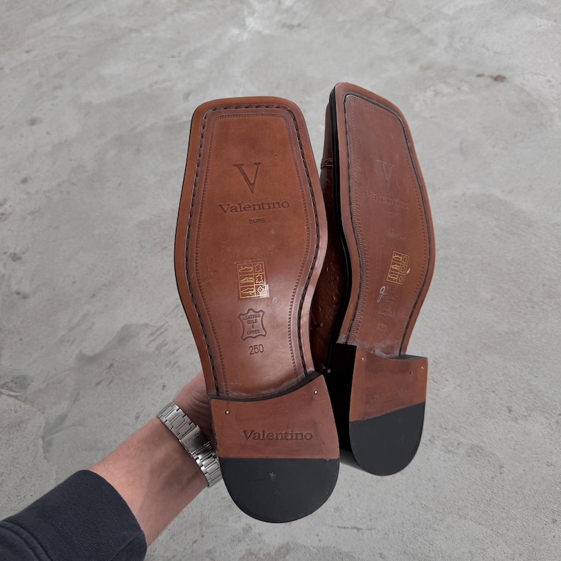 Puede incluir: Un par de zapatos de cuero marr&oacute;n con suela negra. Los zapatos tienen un logotipo V y el texto "Valentino" en la parte posterior. Los zapatos son talla 250.