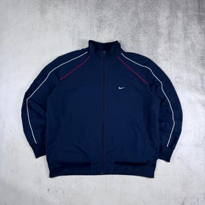 May include: A navy blue track jacket with white and red piping. The jacket has a zipper closure and a small white Nike logo on the left chest.