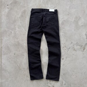 May include: Black denim jeans with a classic five-pocket design. The jeans have a straight leg fit and a small white tag on the back. The pants are displayed on a gray concrete surface.