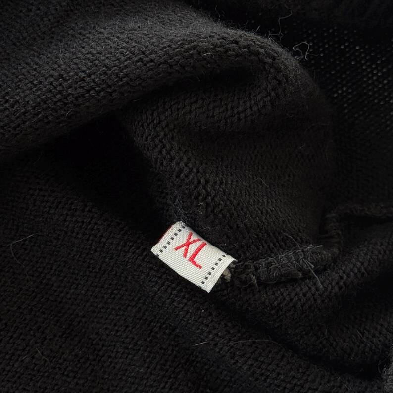 May include: Close-up of black fabric with a small rectangular tag. The tag is white with the letters "XL" in red. The fabric appears to be a knit or woven material.