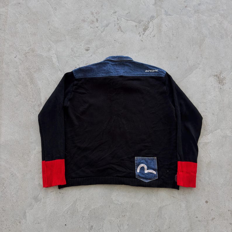 May include: Black sweater with a denim blue upper back and collar. The sleeves have red cuffs. A small denim pocket with a white logo is on the lower right. The word "evisu" is embroidered in white on the upper back.
