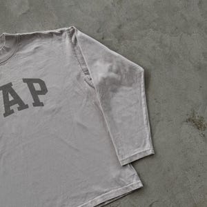 May include: A light gray long-sleeve shirt featuring the word GAP printed in a dark gray block font on the front. The garment has a crew neckline and long sleeves with stitched cuffs, constructed from a solid-colored cotton fabric.