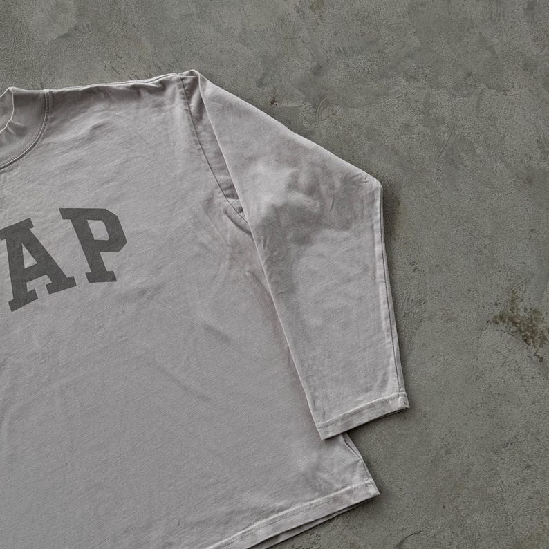 May include: A light gray long-sleeve shirt featuring the word GAP printed in a dark gray block font on the front. The garment has a crew neckline and long sleeves with stitched cuffs, constructed from a solid-colored cotton fabric.