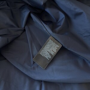 May include: Close-up of a navy blue jacket interior. A black tag with white text is visible, listing the materials and composition of the jacket. The lining is a darker shade of blue.