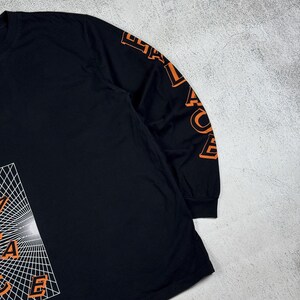 May include: Black long-sleeve shirt with an orange graphic of a geometric design and the word "Palace" on the sleeve.