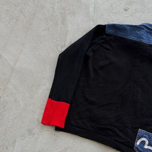 May include: Black long-sleeved shirt with a red cuff detail. The shirt features a denim panel on the upper back and a denim pocket with a white logo. The garment is made of a soft, comfortable material.
