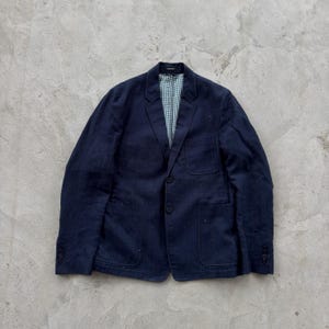 Könnte beinhalten: Ein dunkelblaues Sakko mit dezentem Nadelstreifenmuster, steigendem Revers und zwei Knöpfen auf der Vorderseite. Die Jacke verfügt über eine Brusttasche, zwei aufgesetzte Taschen unten und ein helles Innenfutter mit kleinem grünem Karomuster.
