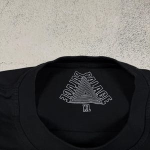 May include: Black t-shirt with a white triangle logo that says "Palace" in a stylized font. The shirt has a size tag that says "XL".
