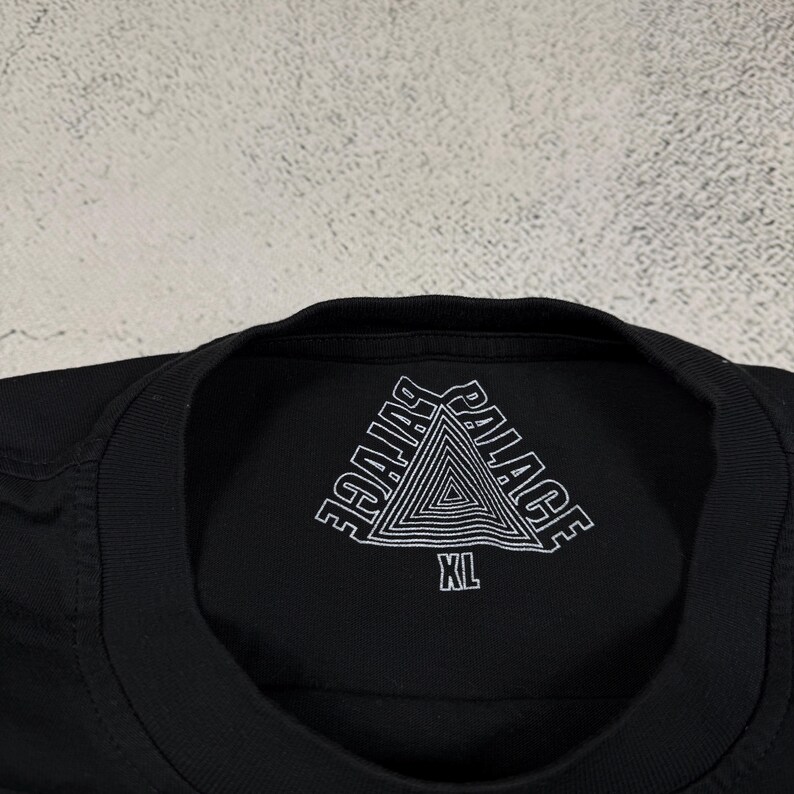 May include: Black t-shirt with a white triangle logo that says "Palace" in a stylized font. The shirt has a size tag that says "XL".