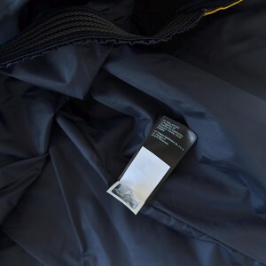May include: Close-up of a navy blue jacket interior with a black label attached. The label has white text and a white blank space. The jacket has a yellow trim and a black, textured collar.