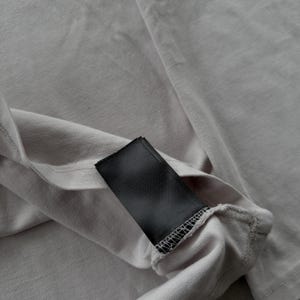 May include: A light gray fabric garment featuring a rectangular black tag sewn into the interior seam. The fabric has a visible knit texture with subtle folds and creases, and the black tag is blank with no visible text or branding printed on it.