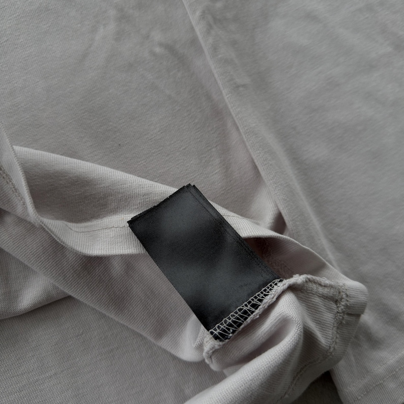 May include: A light gray fabric garment featuring a rectangular black tag sewn into the interior seam. The fabric has a visible knit texture with subtle folds and creases, and the black tag is blank with no visible text or branding printed on it.