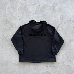 Op de afbeelding: Zwart hoodie met de tekst "ESSENTIALS FEAR OF GOD" gedrukt op de rug.