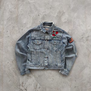 Puede incluir: Chaqueta vaquera azul claro con aspecto desgastado. Presenta bordado de rosas rojas en el pecho y un parche de la bandera americana en la manga. La chaqueta tiene botones, bolsillos y un cuello clásico.