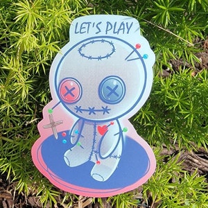 May include: A sticker featuring a cartoon voodoo doll with button eyes, pins, and stitches. The doll is on a blue and pink base, with the words "LET'S PLAY" above it. The sticker is set against a backdrop of green foliage and mulch.