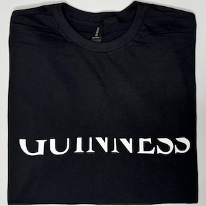 May include: Black t-shirt featuring the word "GUINNESS" in white lettering. The shirt is neatly folded. The Gildan brand label is visible. The t-shirt is displayed on a white surface.