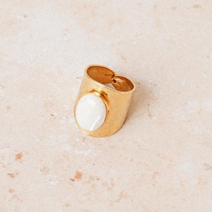Op de afbeelding: Een gouden ring met een witte ovale steen in het midden. De ring is breed en heeft een eenvoudig ontwerp.