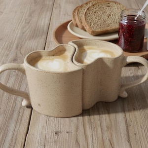 May include: Two beige ceramic mugs, joined together, filled with coffee and topped with foamed milk. The mugs sit on a wooden surface, with a small plate of bread, a jar of jam, and a spoon in the background. The mugs have a unique heart-shaped design.