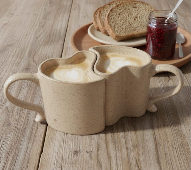May include: Two beige ceramic mugs, joined together, filled with coffee and topped with foamed milk. The mugs sit on a wooden surface, with a small plate of bread, a jar of jam, and a spoon in the background. The mugs have a unique heart-shaped design.