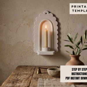 a white candle holder with a lit candle inside, placed on a wooden table. There is also a potted plant and a small bowl on the table. The background is a plain white wall.