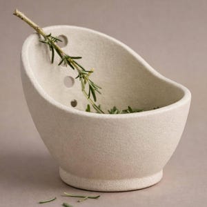 A white ceramic bowl with a curved shape, containing a sprig of green herbs or a similar plant.