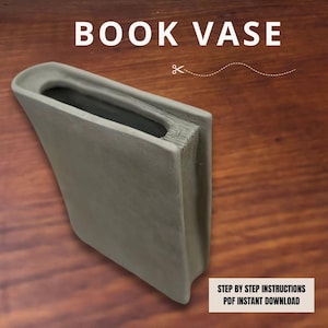 Puede incluir: Un jarrón gris con forma de libro y una abertura rectangular. El jarrón tiene un aspecto texturizado, que se asemeja a las páginas de un libro. El texto "BOOK VASE" se muestra en la parte superior. La esquina inferior derecha tiene el texto "STEP BY STEP INSTRUCTIONS PDF INSTANT DOWNLOAD".