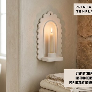 a white scalloped-edge wall-mounted candle holder with a lit candle inside, placed on a shelf next to a stack of towels.